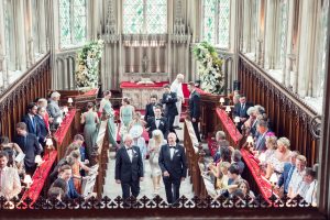 A wedding ceremony in a grand church with ornate stained glass windows. Guests are seated in pews, bridesmaids and groomsmen are walking down the aisle, and the bride and groom stand near the altar with officiants.