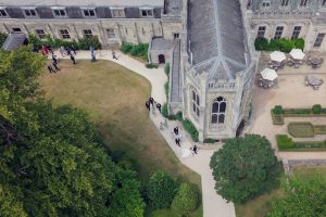 Aerial view of a historic stone building with large windows, surrounded by green lawns, trees, pathways, and people walking and gathering outside. Nearby, tables with parasols and manicured flowerbeds are visible.