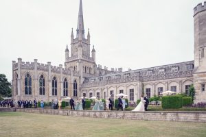 A wedding party, including a bride and groom, walks in a queue outside a large historic stone building with gothic architecture and a tall spire, surrounded by well-kept lawns and gardens.