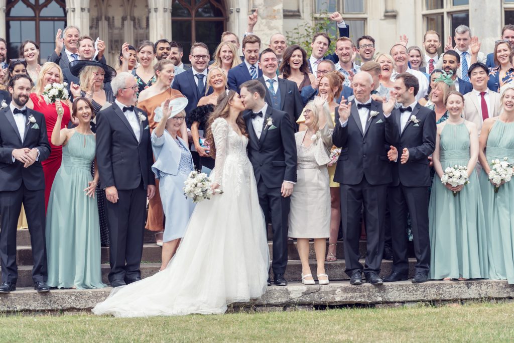 A large wedding party poses outdoors on stone steps. The bride and groom stand in front, kissing, surrounded by bridesmaids in light blue dresses and groomsmen in black suits. Guests behind them are smiling and waving.