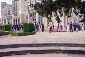 A group of well-dressed people walk and gather outside a grand historic building with large windows and stone arches, likely attending a formal event. Trimmed hedges and a tree branch frame the scene.