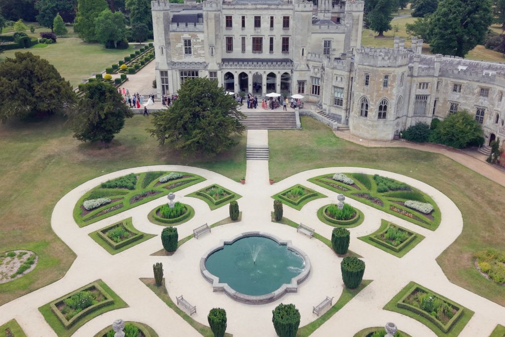 Aerial view of an ornate garden with a central fountain and manicured hedges in front of a large, historic stone mansion surrounded by lawns and trees.