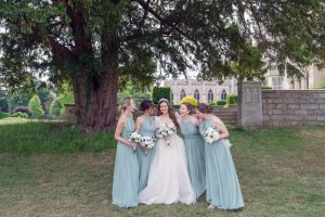 Five women in light blue dresses, one in a white wedding dress, stand together smiling and holding bouquets outdoors near trees and a stone building, suggesting a wedding or bridal party.