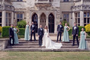 A bride and groom stand in the centre of eight bridesmaids and groomsmen, all dressed formally, posing on stone steps in front of a historic building with large arched windows and detailed stonework.