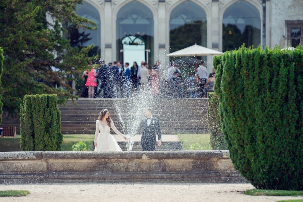 A bride and groom hold hands by a large fountain, with water spraying up between them. In the background, elegantly dressed guests gather near a historic building with arched windows and manicured greenery.