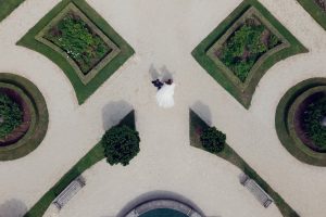Aerial view of a bride and groom walking hand in hand on a symmetrical garden path surrounded by green hedges and flowerbeds.