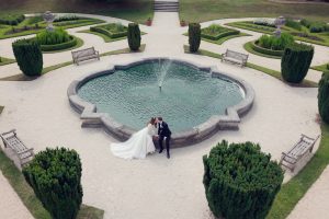 A bride and groom sit beside a decorative fountain in a landscaped garden, sharing a kiss. The scene is viewed from above, showing symmetrical pathways, benches, and manicured greenery surrounding the fountain.