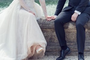 A bride in a white gown and a groom in a black suit sit on a stone ledge, holding hands. The bride’s dress drapes over the side, and both wear wedding rings. Their faces are not visible.