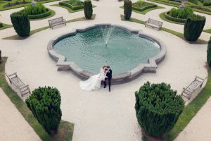 A bride and groom sit together by a decorative fountain in a formal garden, surrounded by green shrubs, benches, and patterned paths, enjoying a quiet moment on their wedding day.