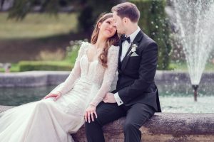 A bride in a white lace gown and a groom in a black tuxedo sit closely together on a stone ledge by a fountain, gazing affectionately at each other. Greenery is visible in the background.