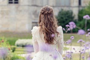 A woman in a white lace wedding dress stands outdoors with her back to the camera, surrounded by purple flowers and greenery. Her long, wavy hair is styled half-up, half-down.