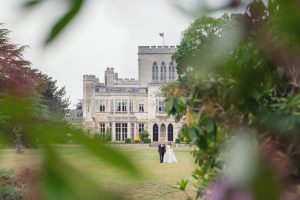 A bride and groom walk hand in hand on a lawn in front of a large, historic stone building, partially framed by green plants and trees in the foreground.