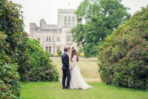 A bride and groom stand hand in hand on a grassy path framed by bushes, looking towards a large, elegant mansion in the background.