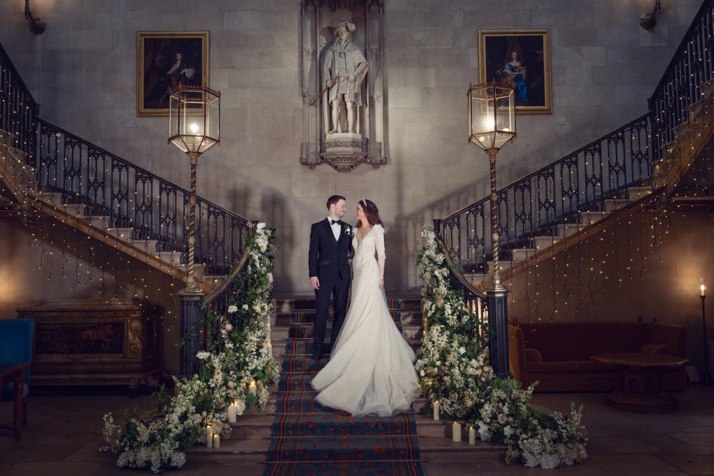A bride and groom stand together on a grand, flower-adorned staircase in a historic hall with stone walls, statues, portraits, and elegant lighting, gazing at each other in wedding attire.