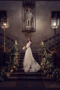 A bride in a long white gown stands on a grand staircase decorated with flowers and candles. She smiles, holding a bouquet, with a statue and ornate lamps in the elegant background.