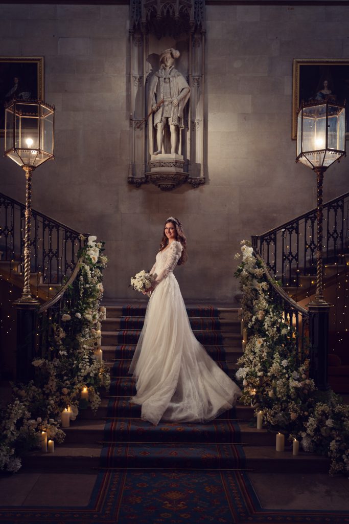 A bride in a long white gown stands on a grand staircase decorated with flowers and candles. She smiles, holding a bouquet, with a statue and ornate lamps in the elegant background.
