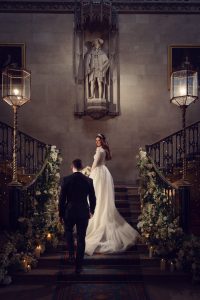 A bride in a long white gown and a groom in a black suit ascend a grand, flower-adorned staircase in an elegant hall, with candles, ornate railings, and a large statue above them.