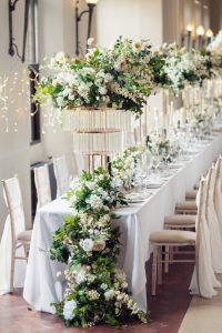 A long banquet table elegantly set with white linen, tall floral centrepieces of white and green flowers, crystal glassware, gold accents, and surrounded by cream-coloured chairs in a bright, airy room.