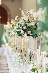 An elegant wedding table is decorated with tall floral centrepieces of white and blush flowers, greenery, and surrounded by lit taper candles and glassware, creating a formal and romantic atmosphere.