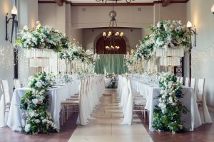 Elegant wedding reception hall with long tables decorated with white tablecloths, tall floral centrepieces featuring white and green flowers, and white chairs. Fairy lights and chandeliers add a romantic atmosphere.