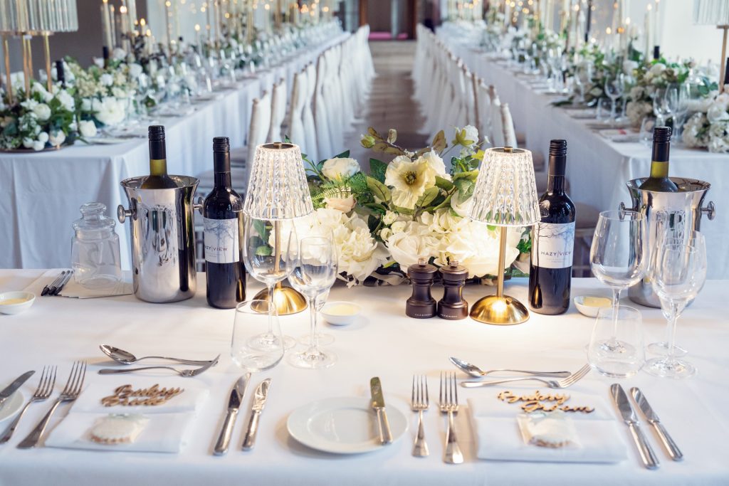 Elegant wedding reception table with white linen, floral centrepieces, wine bottles in coolers, glassware, cutlery, and small lamps, set for a formal meal. Rows of similarly decorated tables extend into the background.