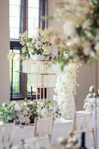 A decorated table set for an elegant event, featuring white flowers, greenery, wine glasses, and a tall centrepiece with hanging glass tubes and floral arrangements, set near large windows.