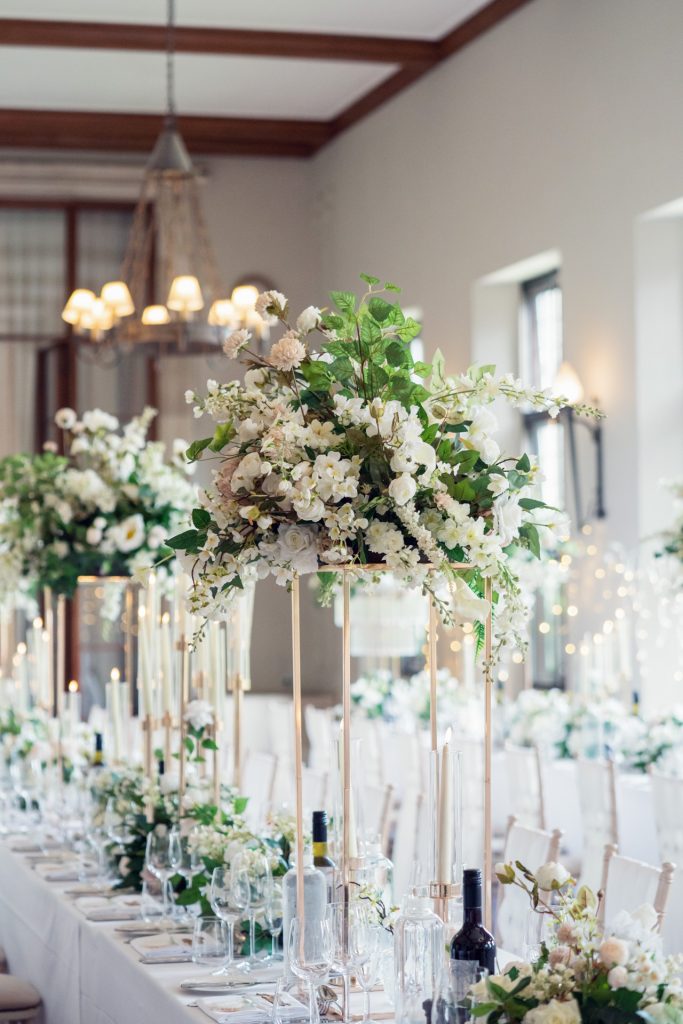 Elegant wedding table setting with tall floral centrepieces of white and green flowers, surrounded by candles, wine bottles, and glassware in a bright, spacious room.