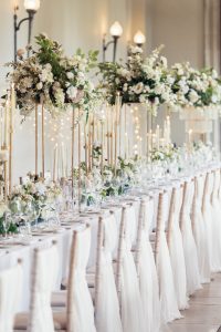 A long, elegant banquet table set for a formal event with white chairs, white linen, tall gold candlesticks, and lush floral centrepieces featuring white and green flowers. Soft lighting creates a romantic atmosphere.