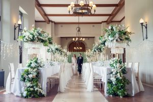 A bride and groom walk down a beautifully decorated wedding aisle lined with elegant white flowers, greenery, and chandeliers in a bright, spacious hall with tall ceilings and white-draped tables.