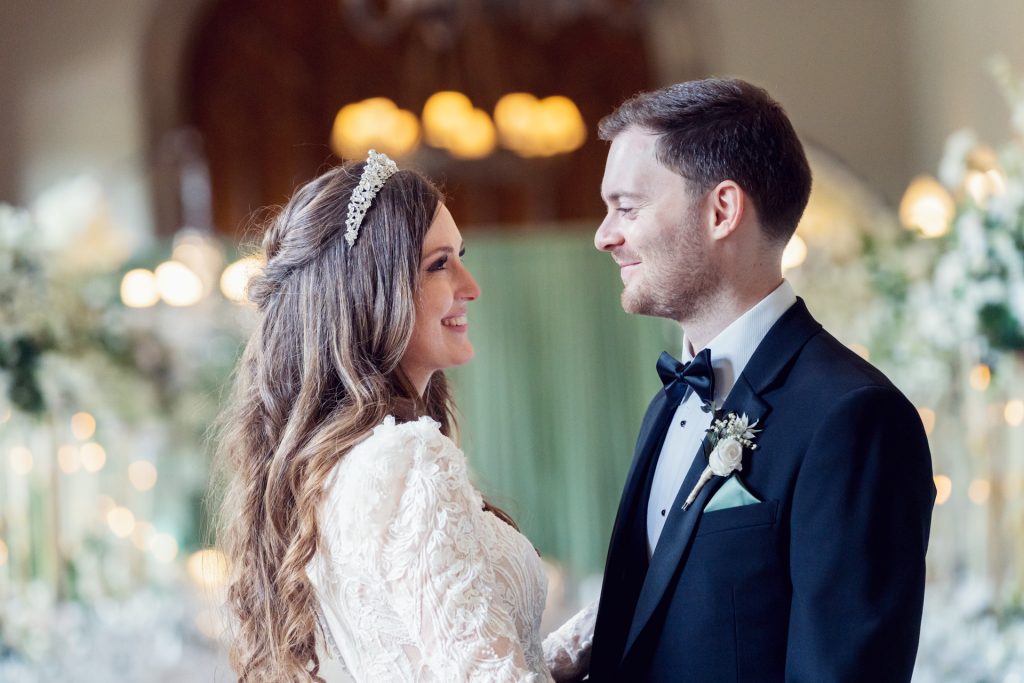 A bride in a white lace dress and tiara smiles at a groom in a black tuxedo. They stand facing each other in an elegant, softly lit venue decorated with flowers.