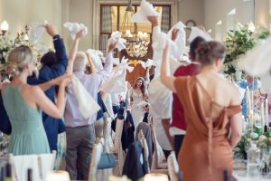 Wedding guests wave serviettes joyfully as a bride and groom enter a decorated reception hall, smiling and holding hands. The scene is lively, with elegant table settings and floral arrangements visible.