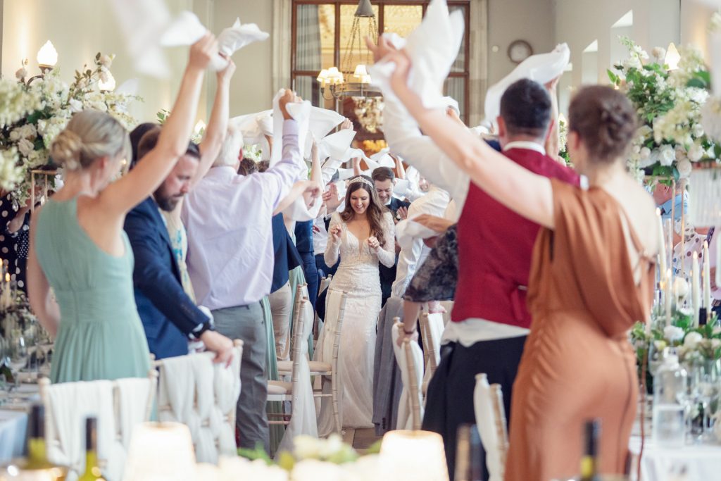 A bride and groom walk down an aisle between dining tables as wedding guests stand, waving serviettes in the air, celebrating their entrance in a bright, decorated reception hall.