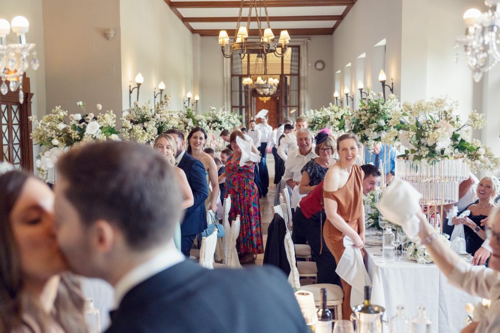 A wedding reception in an elegant, flower-filled hall. Guests stand, waving serviettes and smiling, while a couple in the foreground kiss. The atmosphere is joyful and celebratory.