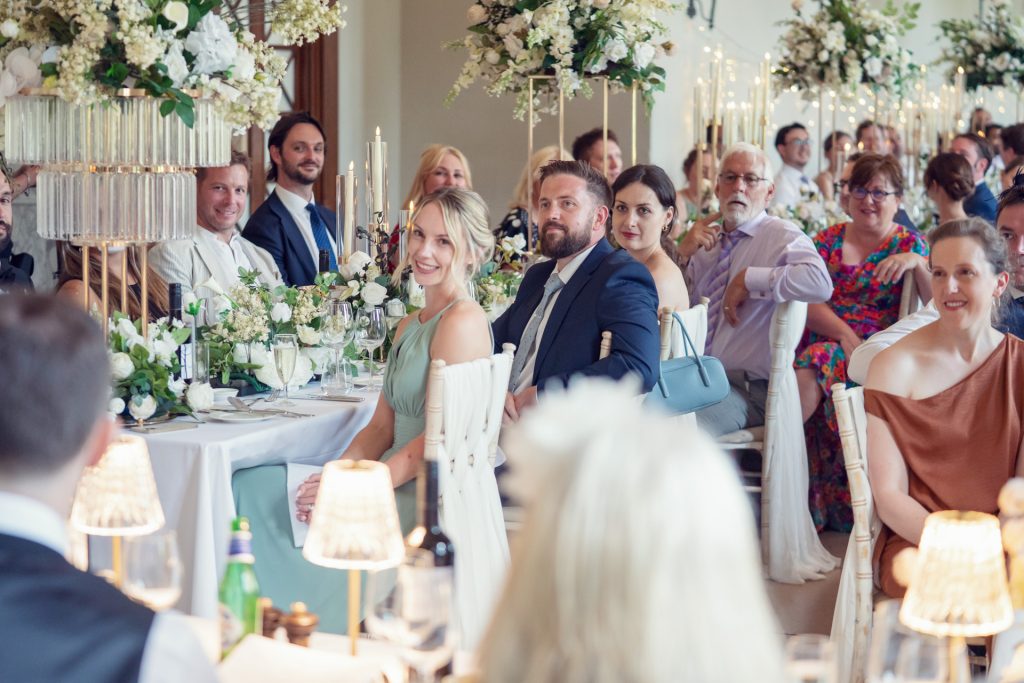 Guests in formal attire sit at elegantly decorated tables with white flowers and candles, smiling and looking towards the front at a wedding or formal event held in a bright, flower-filled room.