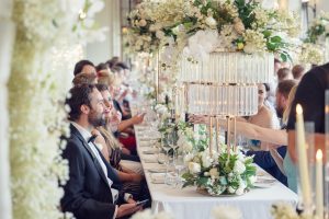 Elegant wedding reception with guests seated at a long, white-decorated table adorned with tall floral centrepieces, candles, and glassware. People are dressed formally, smiling and conversing in a bright, festive setting.