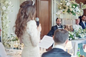 A bride in a white dress gives a speech holding a microphone, while guests, including the groom in a tuxedo, listen at a flower-decorated wedding reception table.