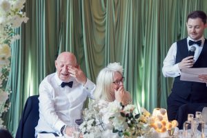 An elderly couple in formal attire laugh and wipe tears from their eyes while seated at a table with flowers, as a man in a waistcoat reads from a paper with a microphone at a celebration.