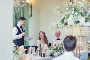 A man in a waistcoat stands and speaks into a microphone, holding a sheet of paper, while a bride in a white dress and a man in a dinner jacket smile at him at a decorated wedding reception table with white flowers.