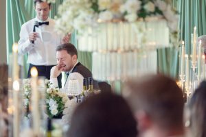 A man in formal wear sits at a table, smiling and touching his face, surrounded by flowers and candles, while another man in a dinner jacket stands nearby holding a microphone and reading from a mobile. The setting is elegant and festive.