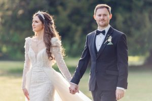 A bride in a long-sleeved lace wedding dress and a groom in a black dinner suit hold hands outdoors, with greenery in the background. The bride looks to the side, while the groom looks ahead.