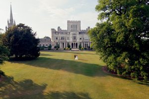 A bride and groom walk hand in hand across a large, green lawn towards a grand historic mansion, surrounded by tall trees and gardens under a partly cloudy sky.