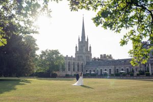 A couple in wedding attire stands on a grassy lawn in front of a large, ornate historic building with a tall spire, framed by leafy green trees and bathed in warm sunlight.