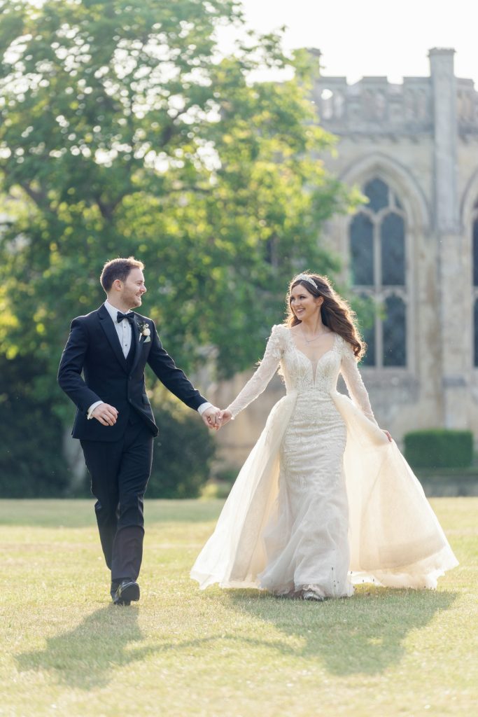 A bride in a long-sleeved white gown and a groom in a black tuxedo walk hand in hand on a sunlit lawn, smiling at each other, with a large tree and a stone building in the background.