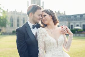 A bride and groom share a tender moment outdoors. The bride wears a white lace gown and tiara, whilst the groom is in a black tuxedo. They smile at each other, holding hands in front of an historic building.