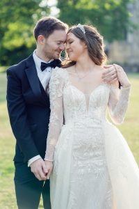 A bride and groom stand close together outdoors, smiling with their eyes closed. The bride wears a lacy white gown and tiara, whilst the groom is in a black tuxedo. They hold hands and touch foreheads, looking happy and in love.