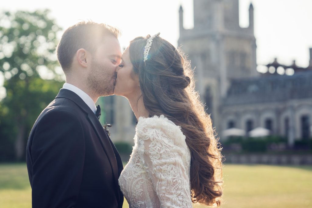 A bride and groom share a gentle kiss outdoors in the sunshine, with a historic stone building in the background. The bride wears a lace dress and headpiece, whilst the groom is in a black suit.