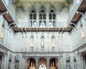 Interior view of a grand hall with Gothic architecture, featuring ornate stone arches, tall windows, decorative columns, and an upper balcony with a metal railing. Light streams in through the arched windows above.