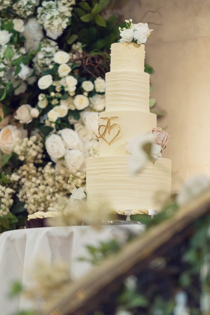 A four-tier white wedding cake decorated with white and blush flowers stands on a table, surrounded by white floral arrangements and greenery in the background.