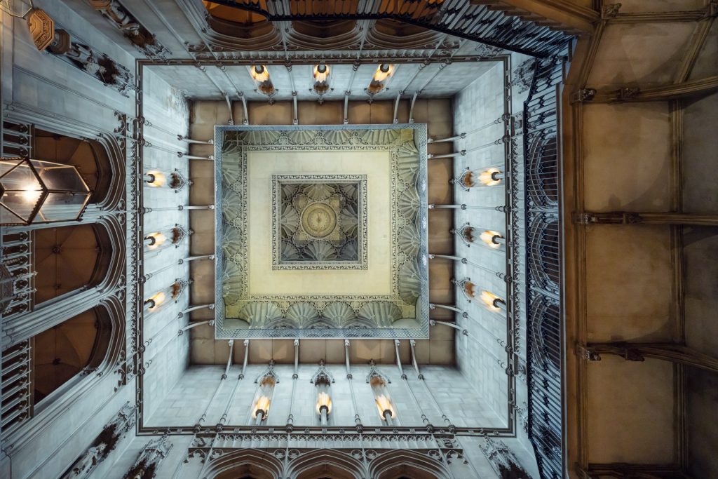 Looking up at an ornate, square ceiling in a grand building, featuring detailed carvings, columns, and glowing wall sconces arranged symmetrically around the central decorative panel.