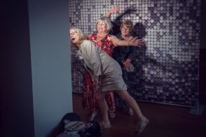 Three older women pose playfully in front of a patterned backdrop, with one peeking round a corner, another stretching her arms upwards, and the third smiling behind them. The mood is fun and light-hearted.
